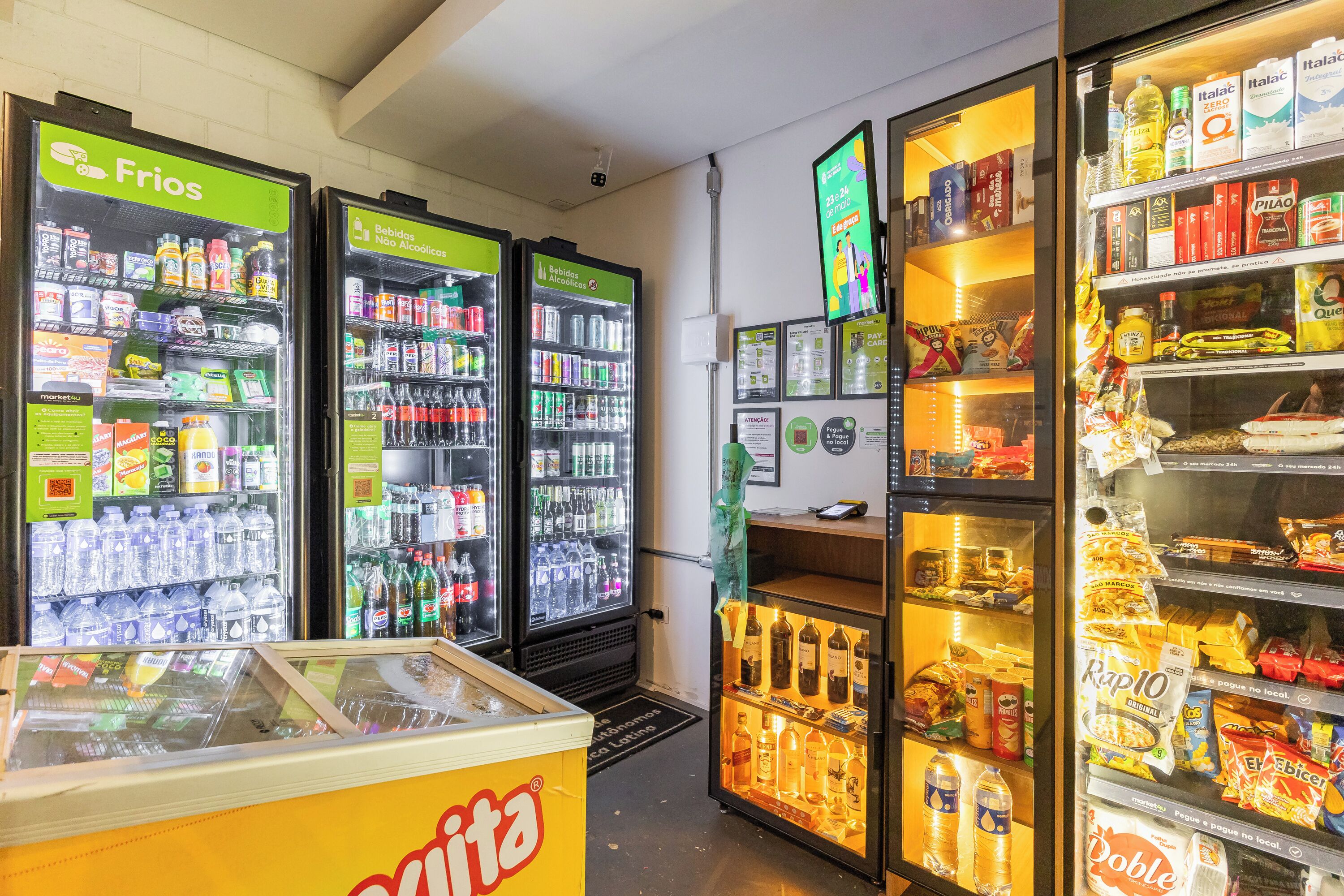Uma loja de conveniência com máquinas de bebidas refrigeradas, prateleiras de lanches e um balcão de caixa.