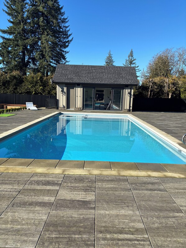 A rectangular swimming pool with a tiled deck, a small building with a dark roof, and a wooden fence in the background.