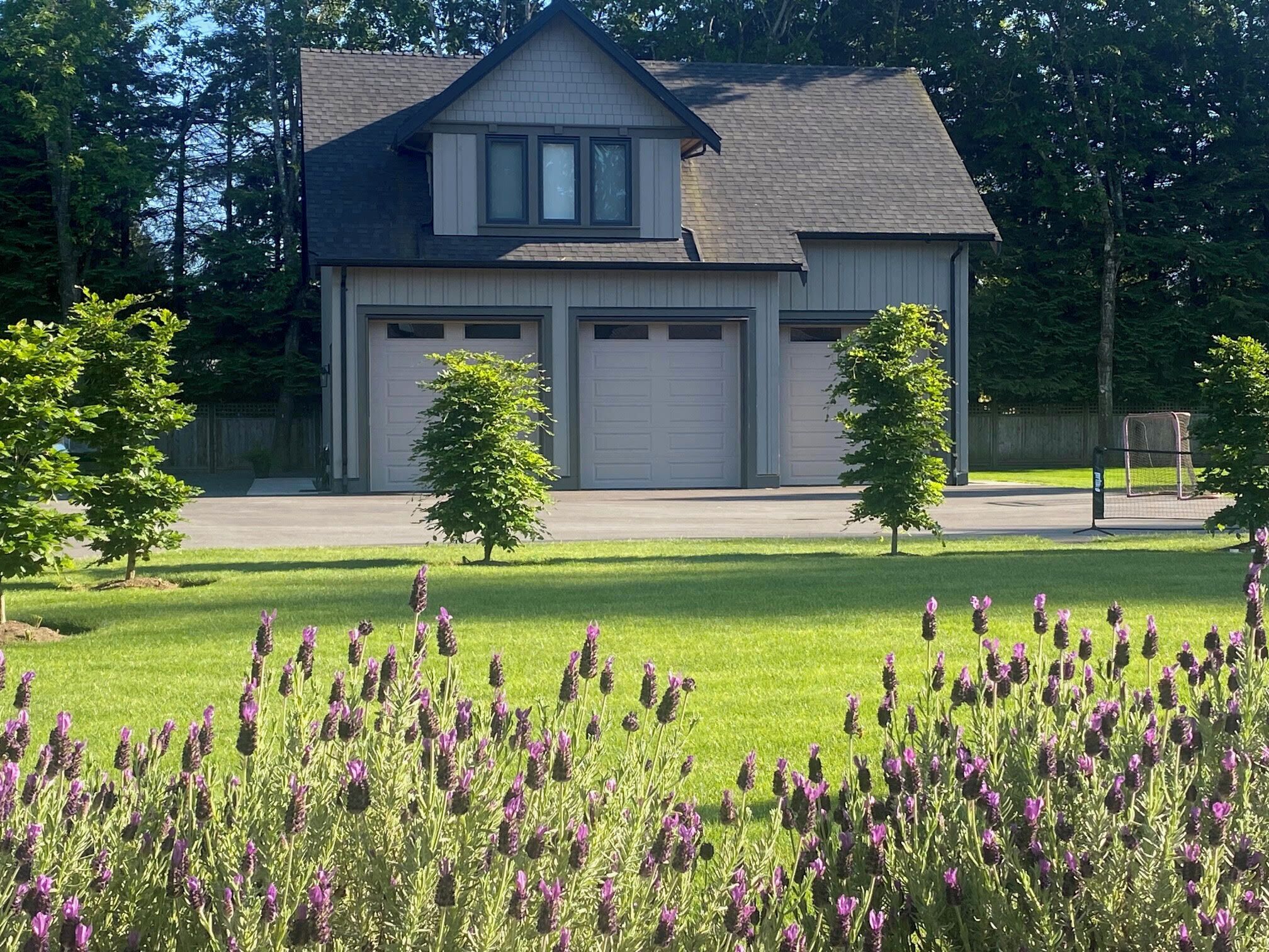 Uma casa de dois andares com garagem, cercada por um gramado bem cuidado e flores roxas.