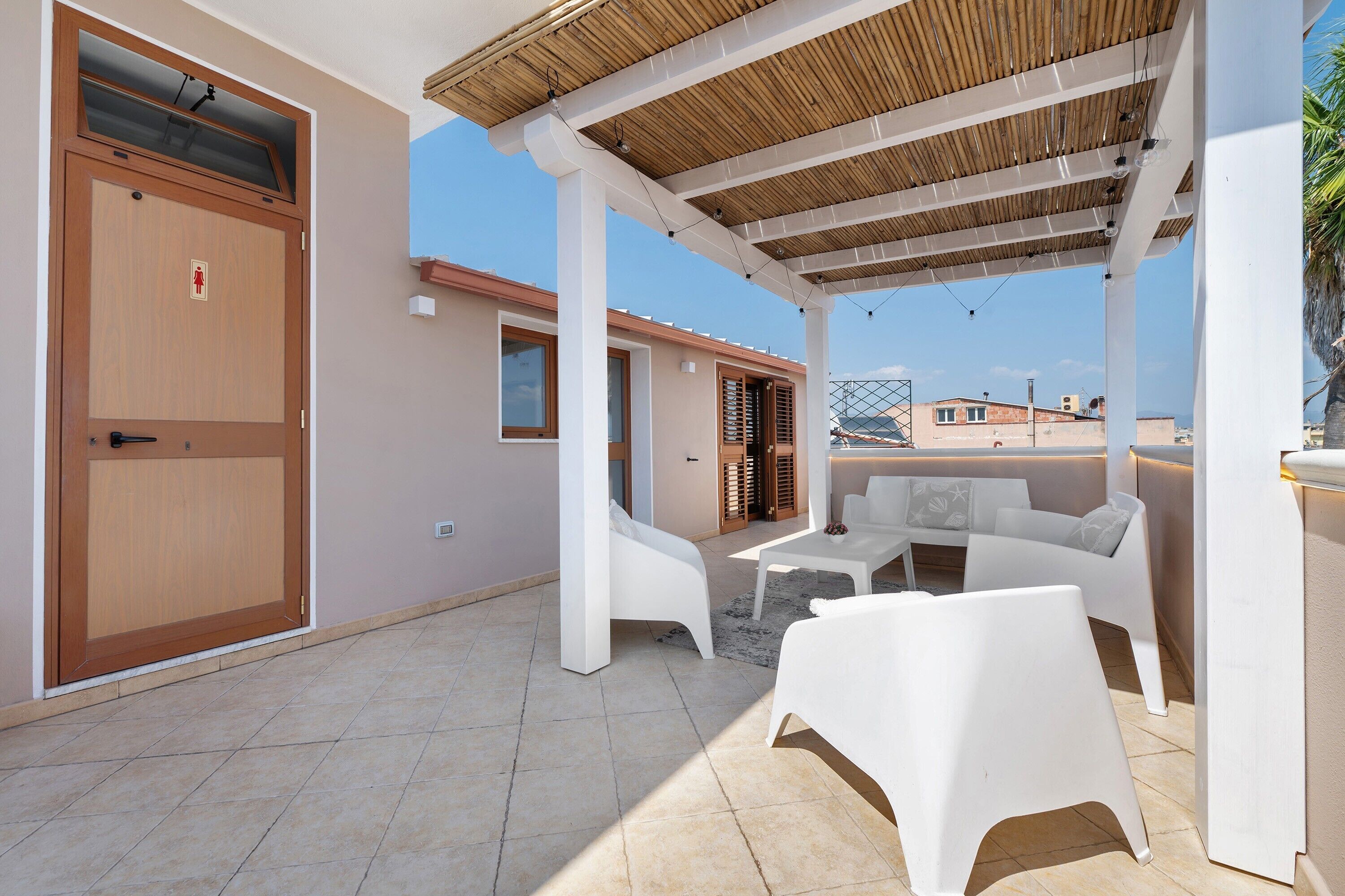 A covered outdoor seating area with white furniture, a wooden door, and a view of buildings in the background.