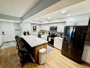 A modern kitchen with a central island, stainless steel appliances, and a white tiled backsplash. - Snowbird Escape: Turnkey 2BR Naples Condo with Golf Views & Screened Lanai (Naples)