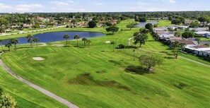 A golf course with a pond, palm trees, and residential buildings in the background. - Snowbird Escape: Turnkey 2BR Naples Condo with Golf Views & Screened Lanai (Naples)