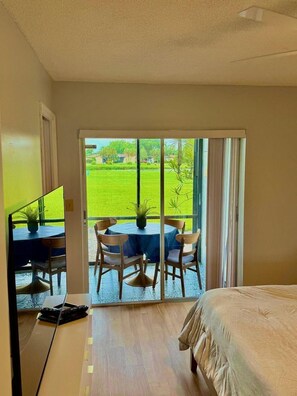 A hotel room with a bed, a desk with a computer, a table with chairs, and a sliding glass door leading to a balcony with a view of a green lawn and trees. - Snowbird Escape: Turnkey 2BR Naples Condo with Golf Views & Screened Lanai (Naples)