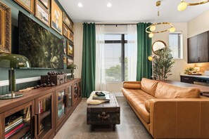 A living room with a brown leather sofa, a wooden cabinet with glass doors, a green curtain, and a large painting on the wall. - Library Retreat | Antiques & Gallery Wall (Nashville)