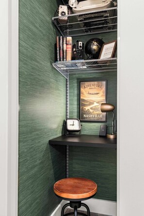 A corner of a room with a green textured wall, a metal shelf with books and a vintage camera, a framed poster of a plane, a clock, a desk lamp, and a wooden stool. - Library Retreat | Antiques & Gallery Wall (Nashville)