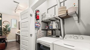A laundry room with a front-loading washer and dryer, a fire extinguisher, a first aid kit, and shelves with towels and cleaning supplies. - Library Retreat | Antiques & Gallery Wall (Nashville)
