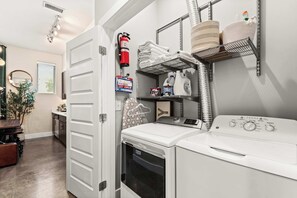 A laundry room with a front-loading washer and dryer, a fire extinguisher, a first aid kit, and shelves with towels and cleaning supplies. - Library Retreat | Antiques & Gallery Wall (Nashville)