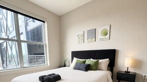 Ein Schlafzimmer mit einem großen Bett, einem Nachttisch, einer Lampe und einem Fenster mit Blick auf Gebäude.