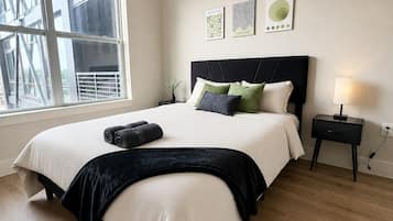 Une chambre à coucher avec un grand lit, une table de chevet, une lampe et une fenêtre donnant sur des immeubles.