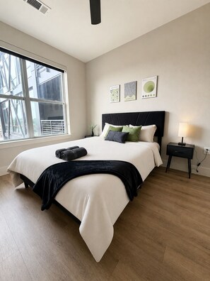 A bedroom with a large bed, a nightstand, a lamp, and a window with a view of buildings. - Skyline Balcony Retreat | Luxury Apartment | Pool (Dallas)