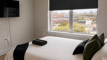 A bedroom with a bed, a TV mounted on the wall, a window with a view of buildings, and a ceiling fan.