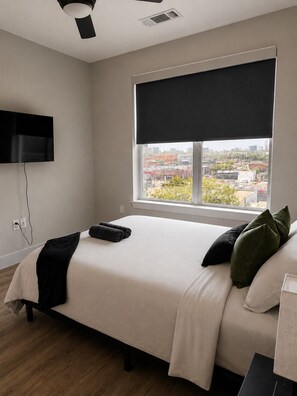 A bedroom with a bed, a TV mounted on the wall, a window with a view of buildings, and a ceiling fan.