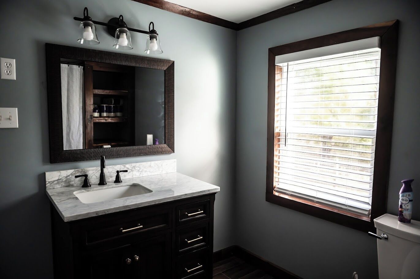 Une salle de bain avec un lavabo en marbre, des meubles sombres, un grand miroir et une fenêtre avec des stores.