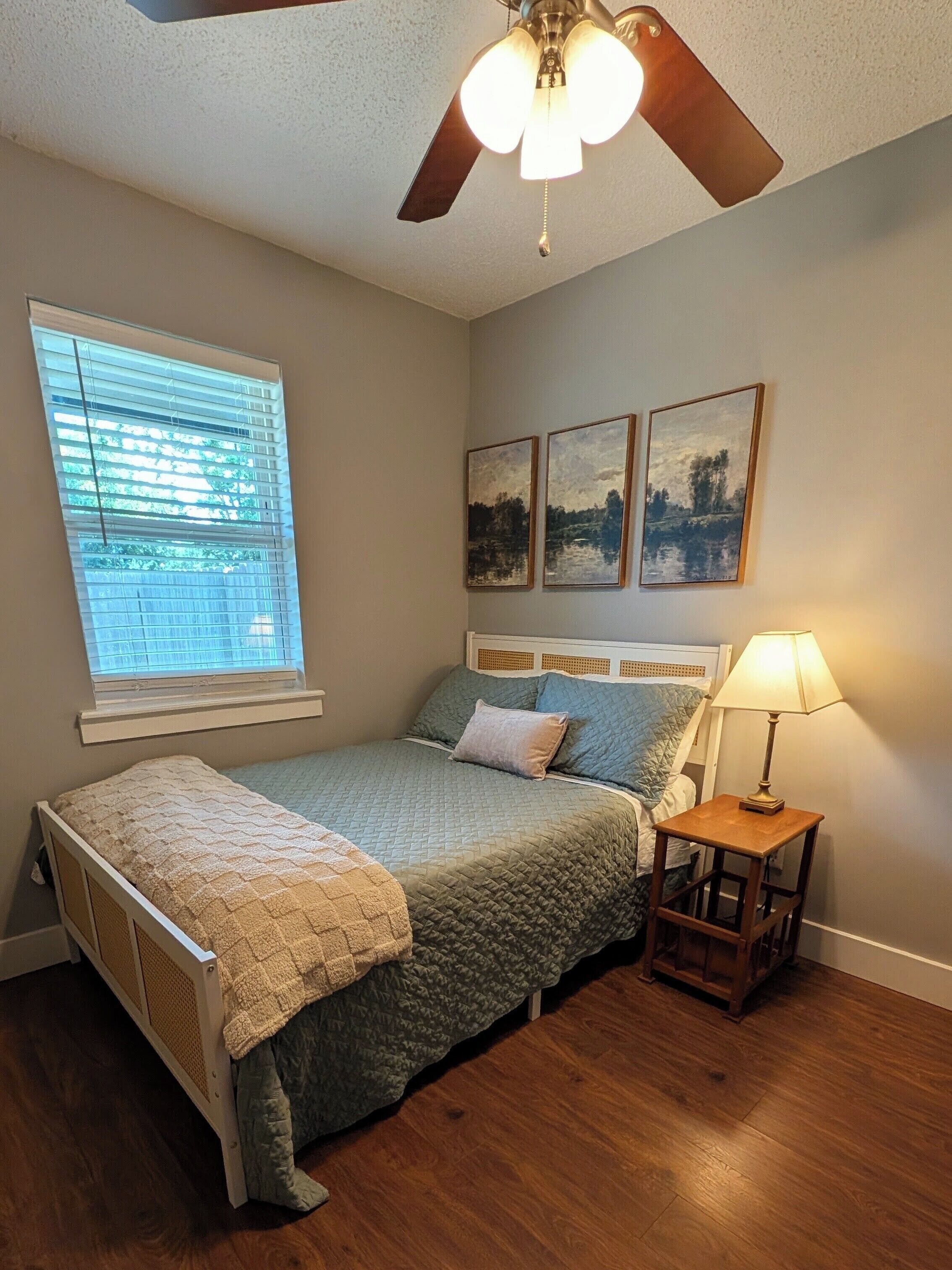 A bedroom with a bed, a nightstand, a lamp, a ceiling fan, and a window with blinds.
