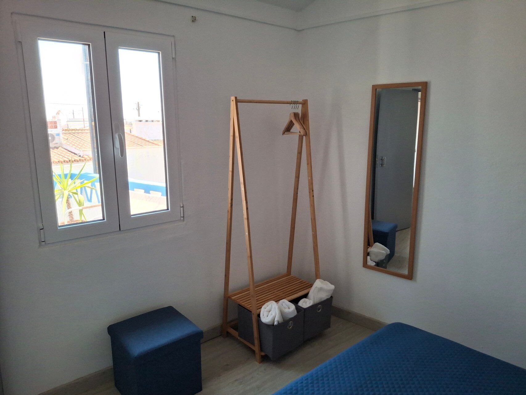 A room with a bed, a hanging chair, a small table, a mirror, and a window with a view of a building and palm trees.