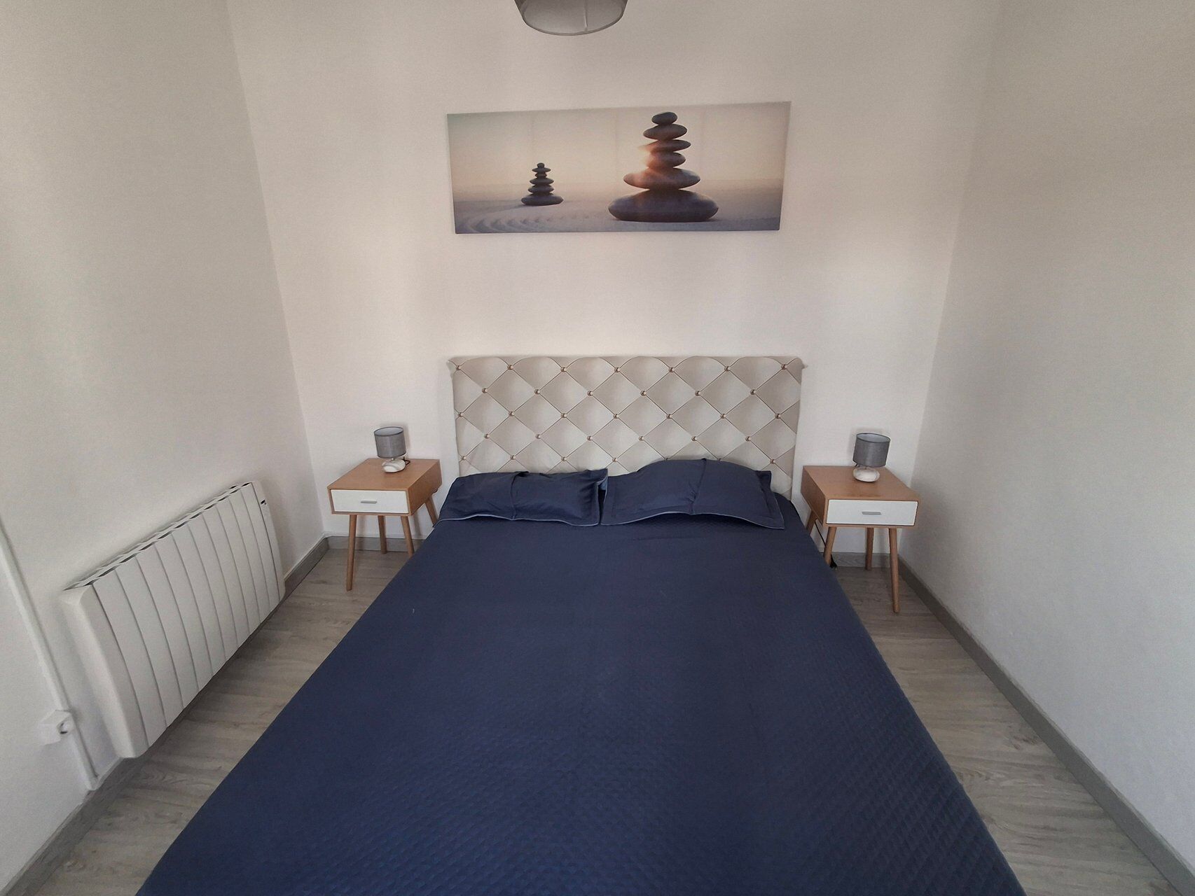 A bedroom with a bed, two bedside tables, a radiator, and a painting of stacked stones.