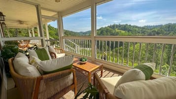 A covered porch with wicker furniture, a small table, and a view of a forested landscape.