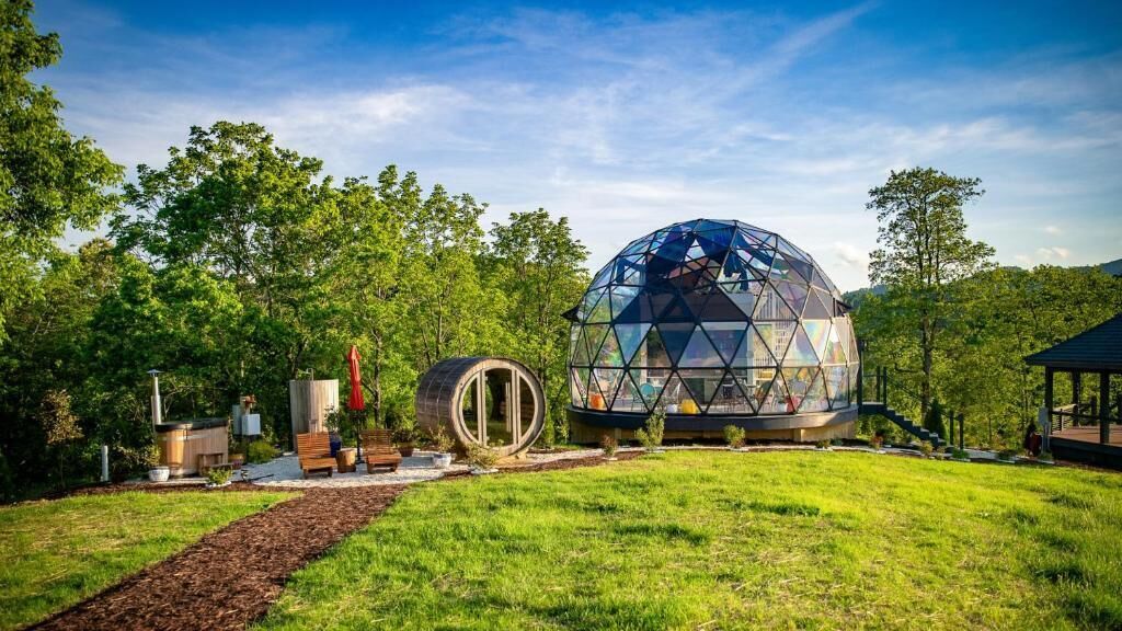 Estructura en forma de domo geodésico con techo transparente, rodeada por un jardín ajardinado con árboles y un cielo despejado.