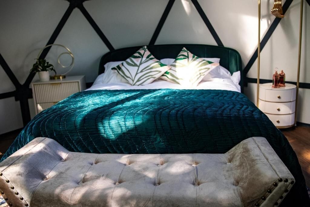 Un lit avec une couette verte, un oreiller blanc et un pouf capitonné.