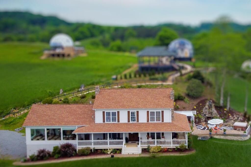 Modelo de una casa con porche, rodeada de un paisaje verde y dos estructuras abovedadas al fondo.