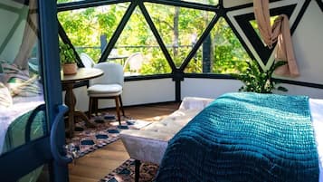 A room with a bed, a chair, a small table, and a view of greenery through a geometric window design.