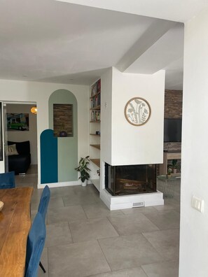 Ein modernes Wohnzimmer mit Kamin, Bücherregal und Essbereich mit Holztisch und blauen Stühlen.