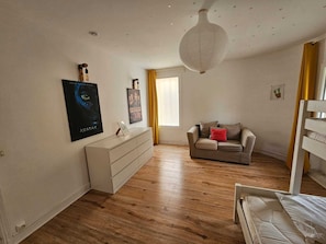 Une pièce comprenant un canapé, une commode et un lit. Il y a une fenêtre avec des rideaux, un luminaire fixé au mur et des affiches sur le mur.