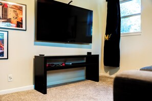 A room with a flat-screen TV mounted on the wall, a black entertainment unit below it, and a window with a curtain. - 5 Min to Downtown - Huge Yard - Dog Friendly -3GB/s Wifi - 75" TV (Austin)