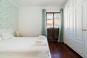 A hotel room with a bed, a nightstand, a window with curtains, and a view of buildings outside. - GuestReady - Dream Corner (Caniço)