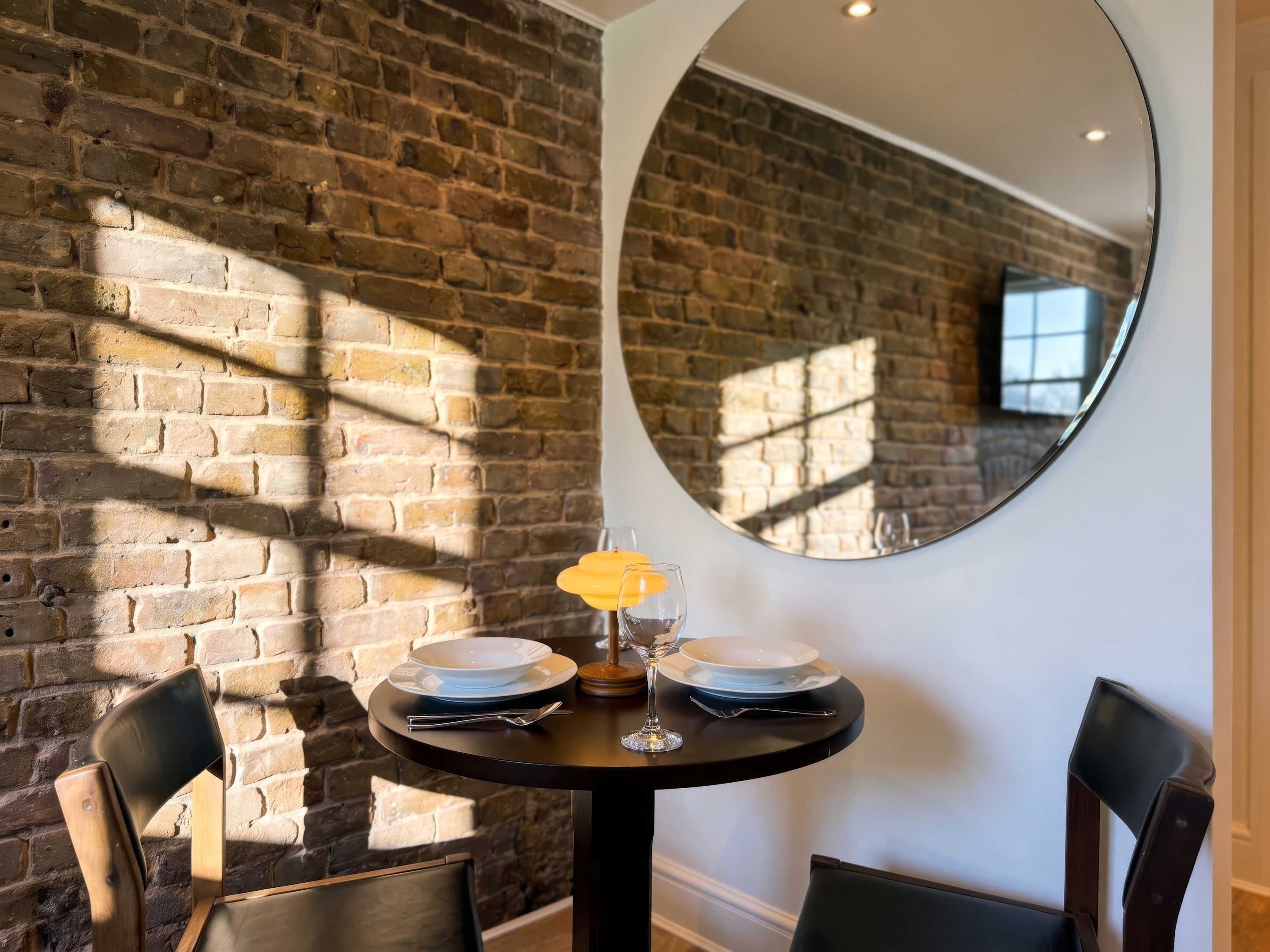 A dining area with a brick wall, a round mirror, and a table set with plates, glasses, and cutlery.