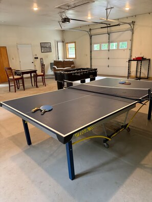 A ping pong table with a table tennis paddle, a sofa, and a projector in a room with a garage door and windows. - Welcome to Bud and Jacquie Sue’s (Sterling)
