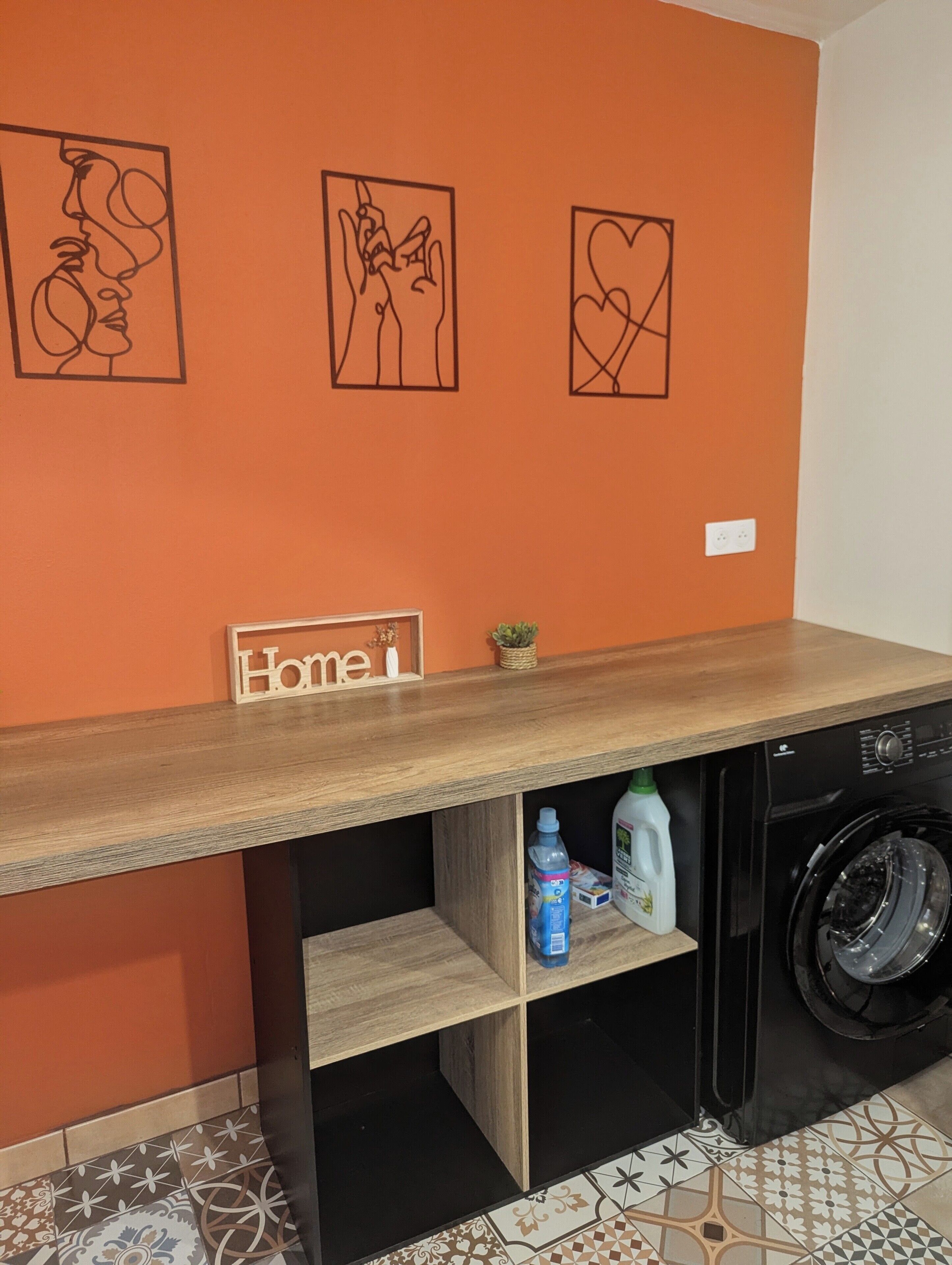 Un espace buanderie comprenant un lave-linge, une étagère en bois avec des produits de nettoyage et une pancarte indiquant « Accueil ».