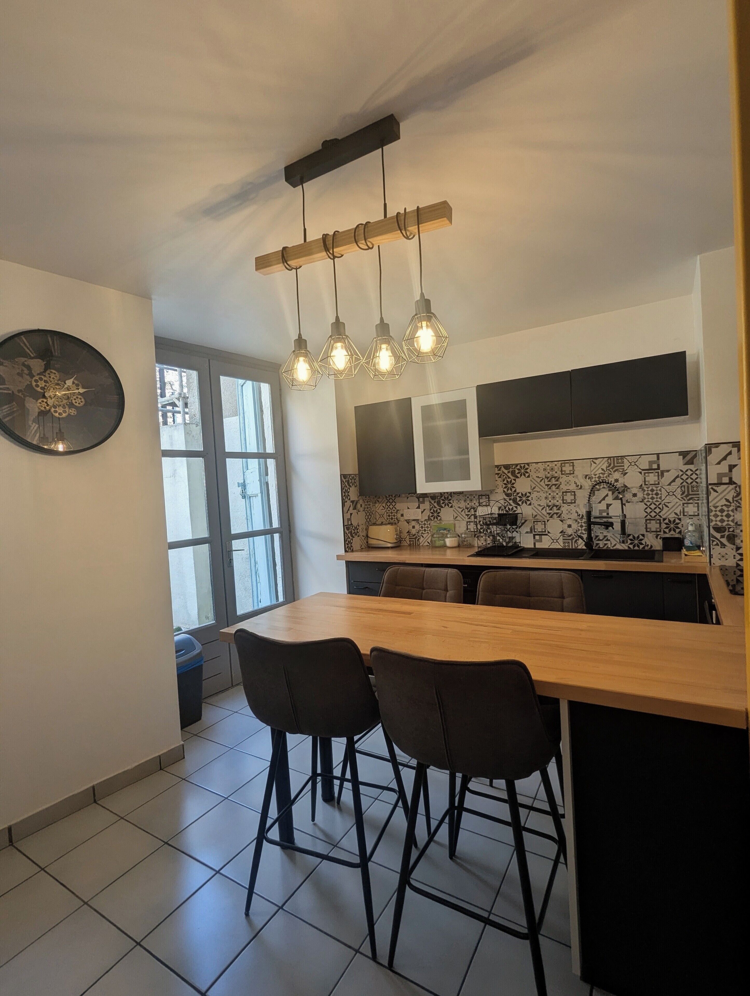 Une cuisine moderne avec un îlot central en bois, des tabourets de bar noirs, une grande horloge murale et des luminaires suspendus.
