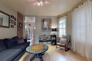 A living room with a sofa, a round coffee table, a bookshelf, a chair, and a window with curtains. - Trendy suite in Mission! Steps away from 17th ave, the river and Stampede (Calgary)