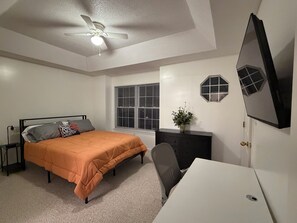 Ein Schlafzimmer mit einem Bett, einem Schreibtisch, einem Stuhl, einem Deckenventilator, einem Fenster mit Jalousien und einem an der Wand befestigten Fernseher.