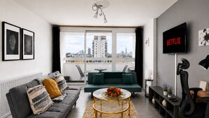 Un salon moderne avec un canapé gris, un canapé vert, une table basse, un téléviseur et une vue sur la ville par la fenêtre.