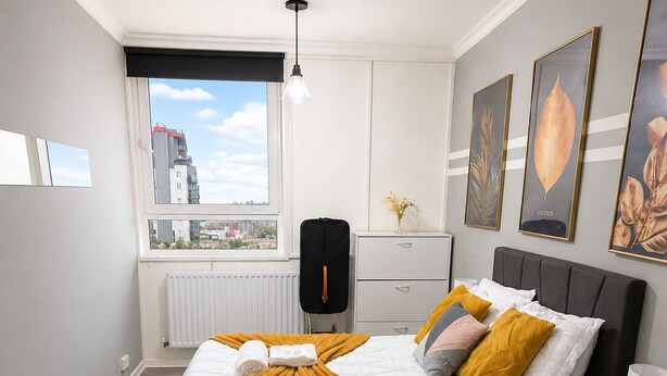 Une chambre moderne avec un grand lit, une vue sur la ville par la fenêtre, un radiateur et un tapis à motifs.