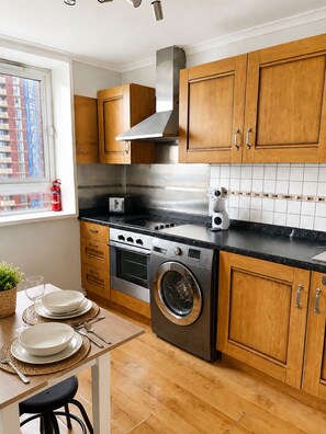 A modern kitchen with wooden cabinets, stainless steel appliances, and a window with a view of buildings. - Canary Wharf Apartment | Thames Views, Balcony & Workspace | Sleeps 6 (London)