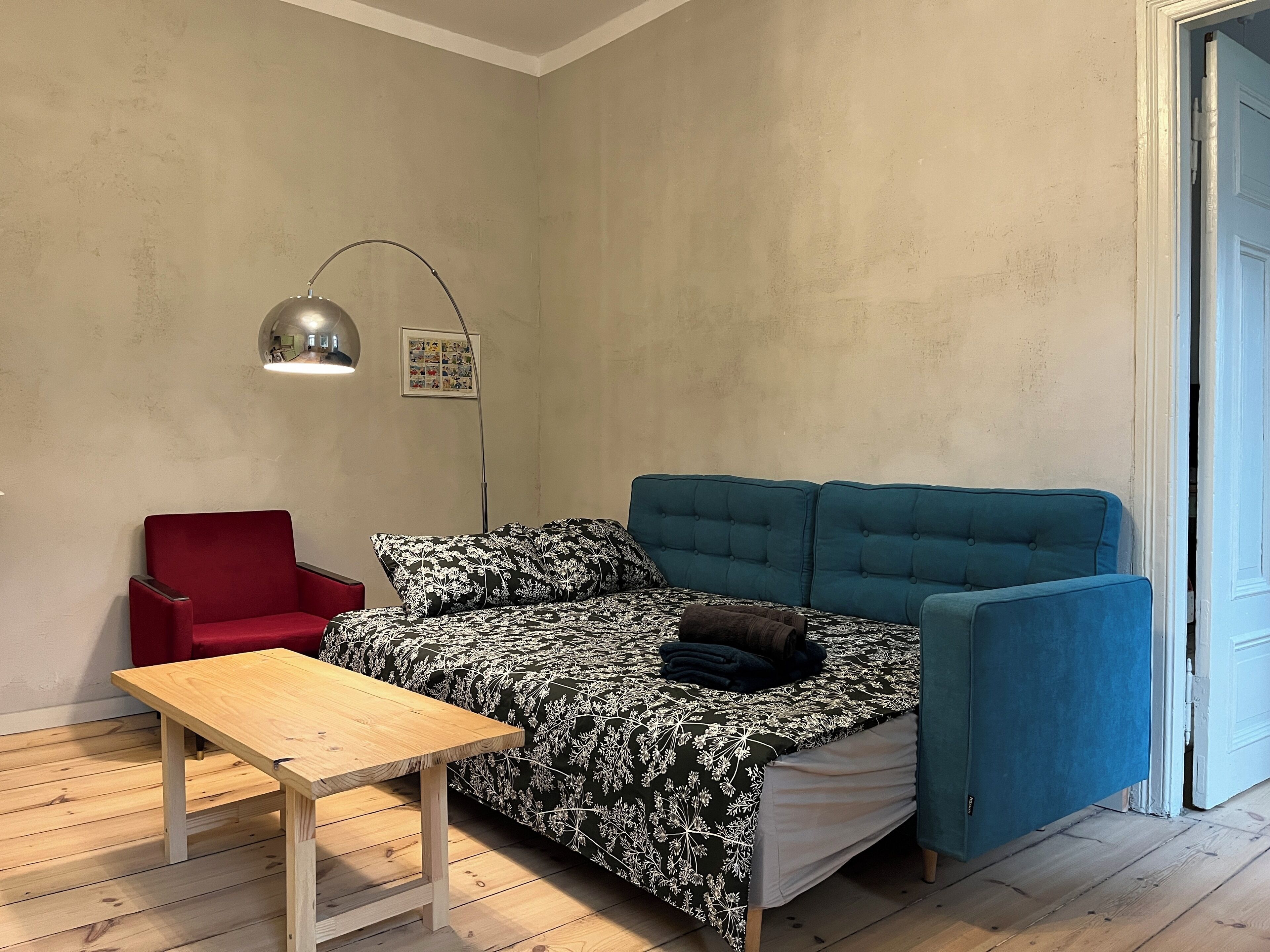 Ein kleines Zimmer mit einem Bett, einem roten Sessel, einem Holztisch und einer Stehlampe.