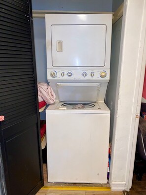 A compact, white washing machine with a front-loading door, situated in a small laundry room with a wooden floor. - Cozy 1-bedroom house in peaceful Los Angeles with AC (Los Angeles)
