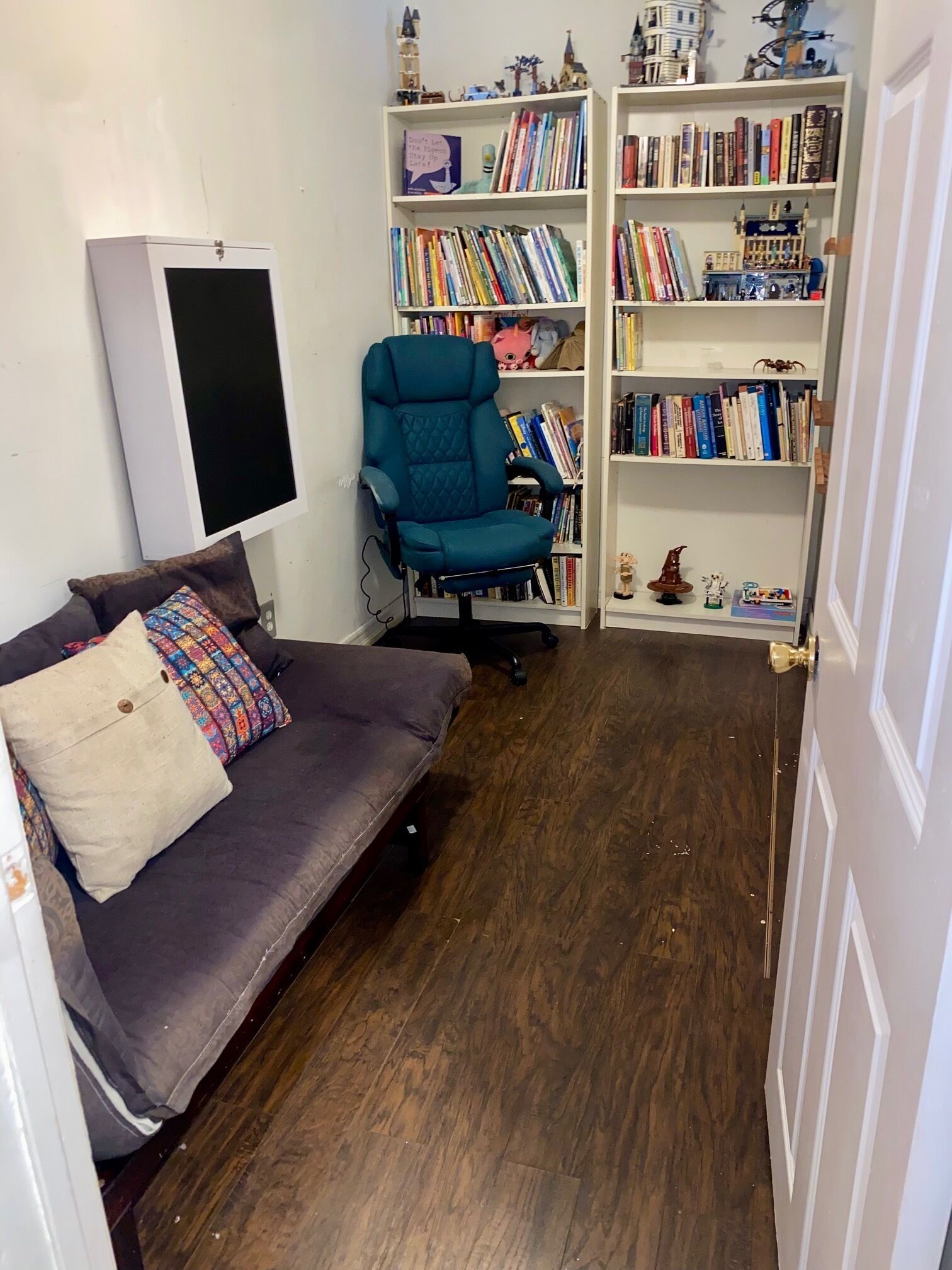 A room with a dark wood floor, a grey sofa with patterned cushions, a teal office chair, and a bookshelf filled with books and figurines.