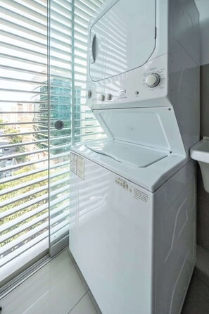A front view of a modern, white laundry room with a large window and blinds, a built-in washer and dryer, and a view of the cityscape outside. - 3 APTS in One Building · Ideal for Large Groups! (Miraflores)