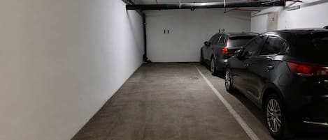 A parking garage with concrete floors, marked parking spaces, and visible pipes and ducts on the ceiling.
