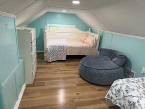 Una habitación pequeña y acogedora con techo inclinado, una cama con ropa de cama floral, un sillón de peluche gris y una mesita con mantel floral.