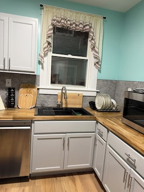 Una cocina con armarios blancos, un lavamanos de acero inoxidable, una tabla de cortar de madera y un escurreplatos.