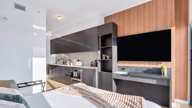 Moderni hotellihuone, jossa on sänky, seinälle asennettu taulutelevisio, sisäänrakennettu keittotila ja oleskelualue.