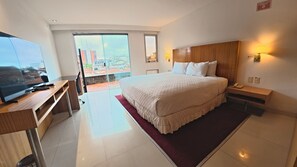 A hotel room with a large bed, a desk with a television, a nightstand, and a view of the city through the window. - Hotel Pascana centro (Santa Cruz)