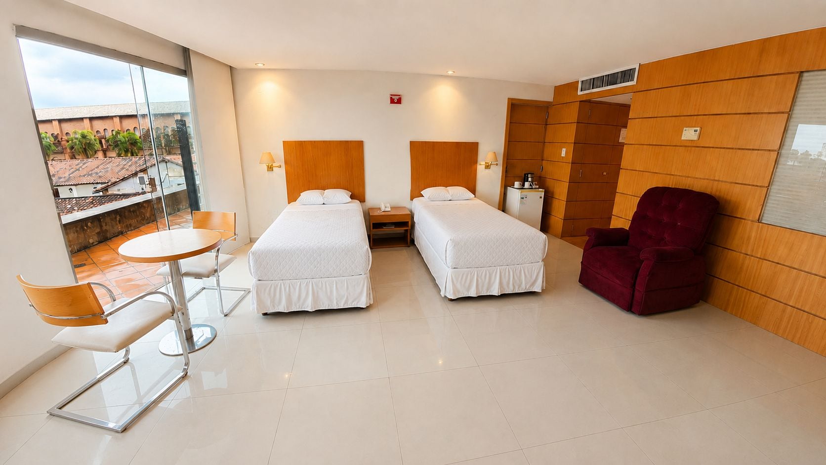 Une chambre d’hôtel avec deux lits, une chaise rouge, une petite table et une vue sur les bâtiments environnants.
