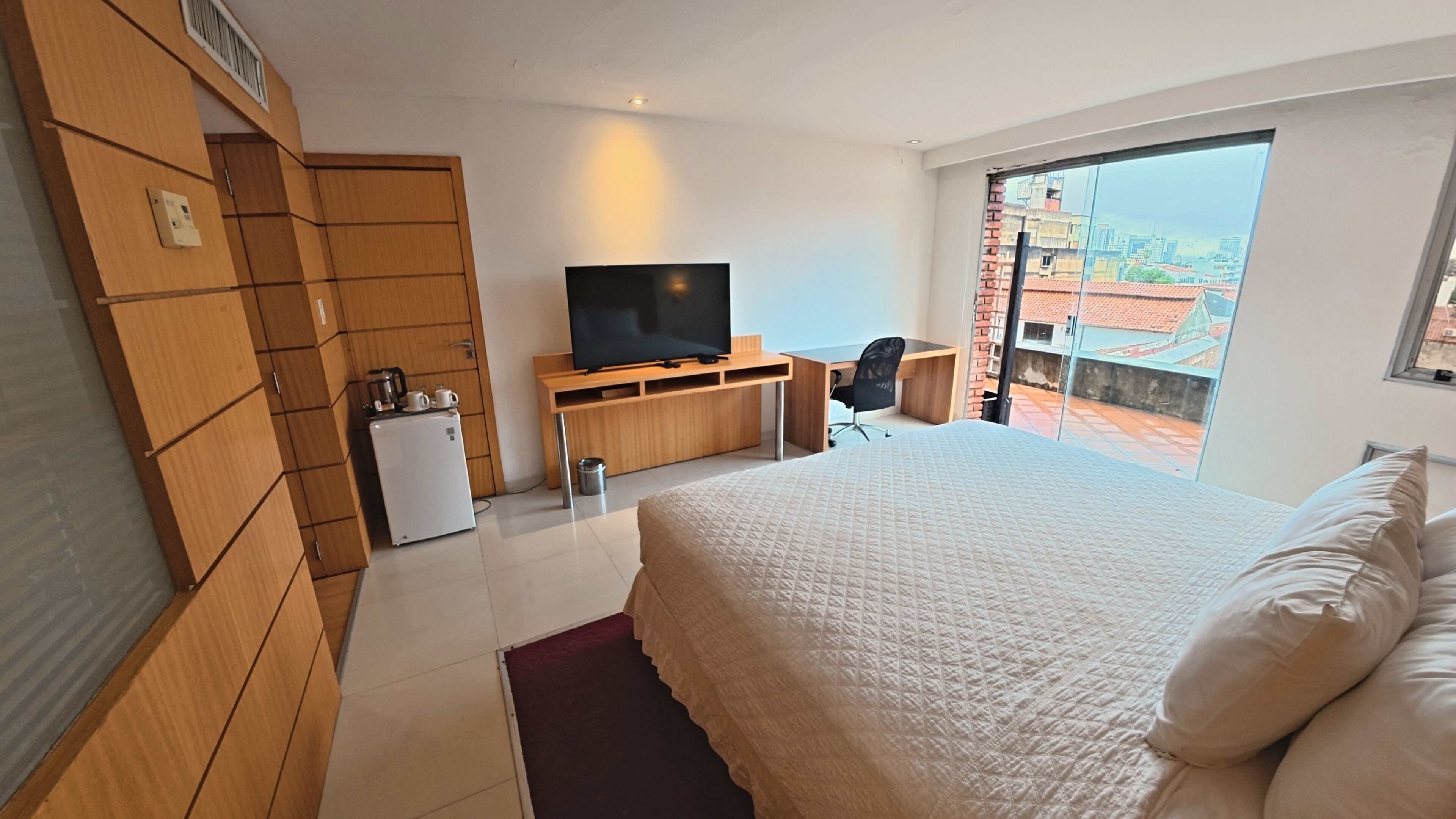 A hotel room with a bed, a TV, a desk, and a view of the city.