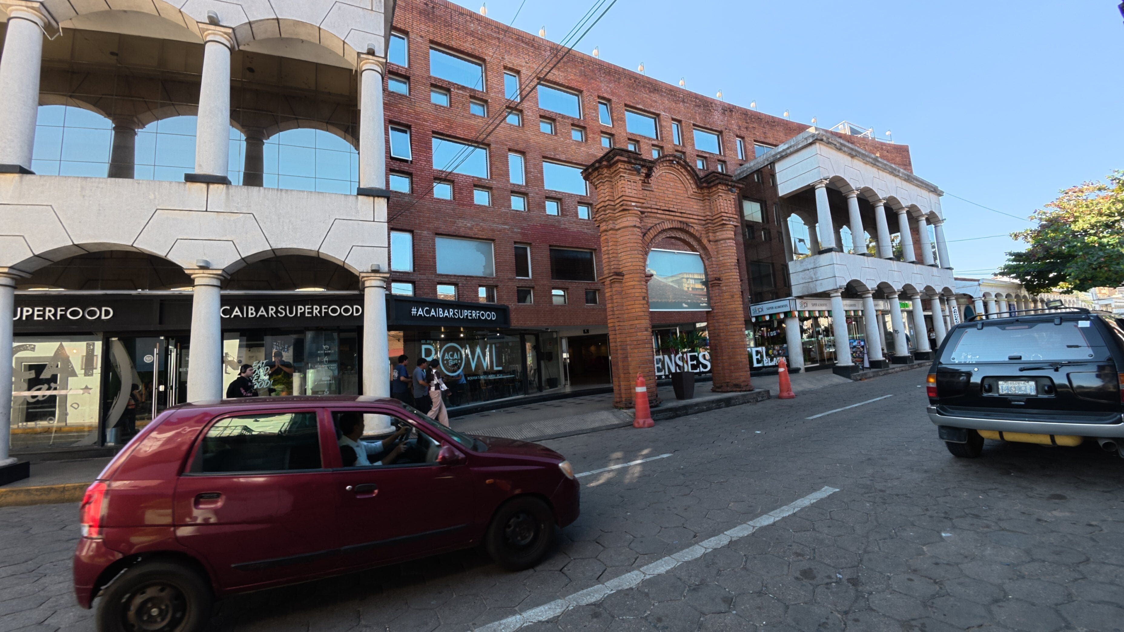 Vista de la calle con un auto rojo estacionado al lado, edificios con escaparates y cielo despejado.